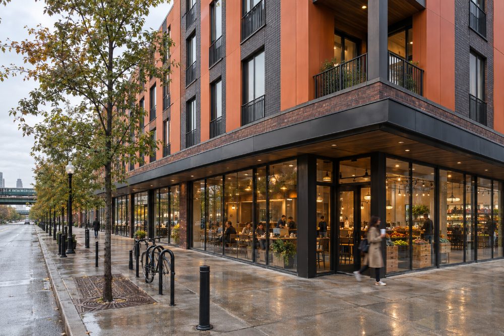 The street frontage on a wet evening, with the caf&eacute; and the grocer lit behind full-height glass and bike racks at the kerb.